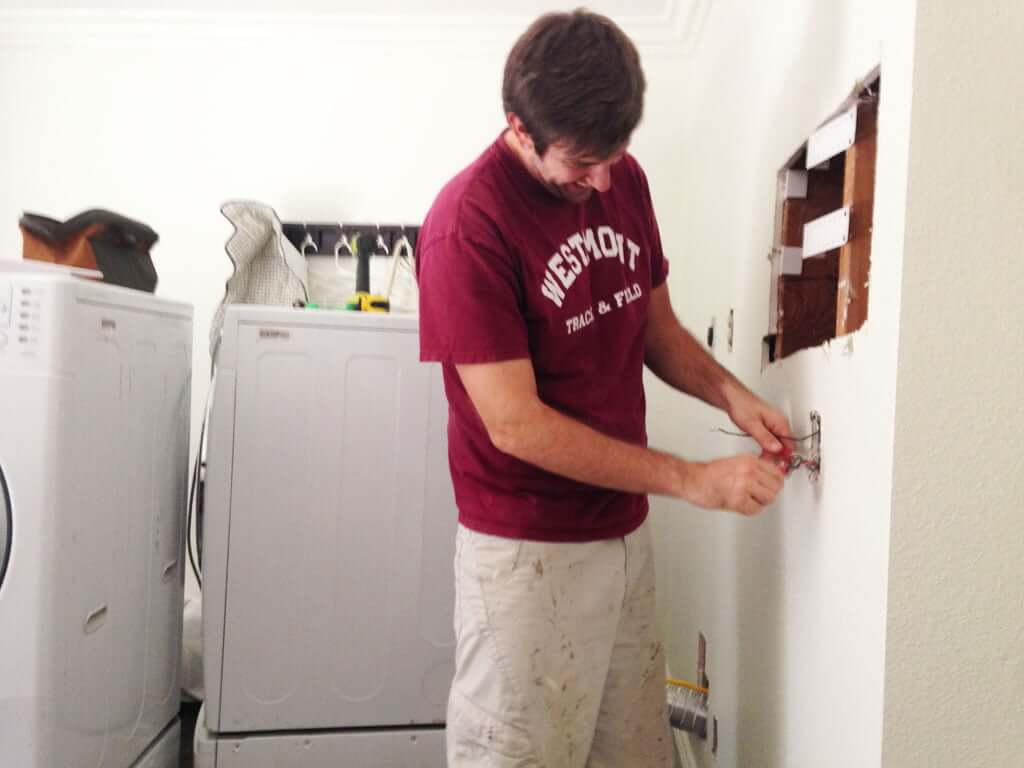 man working on electrical outlet in wall with washer and dryer behind him