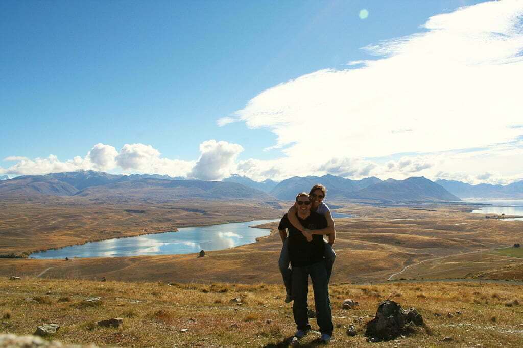 couple in New Zealand with mountains and lakes and grassy hills in distance