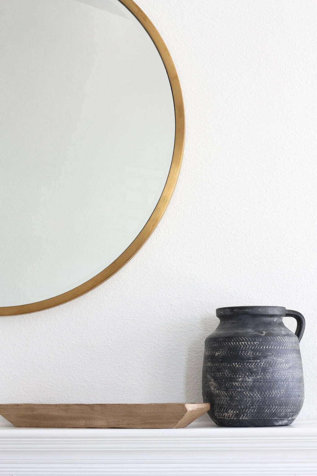 white fireplace mantel with round brass mirror, black clay pot and wood tray