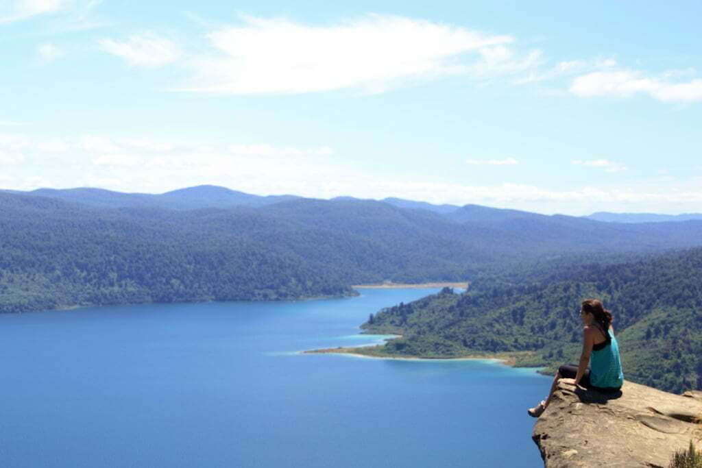 woman sitting on cliff edge overlooking a lake and forest in New Zealand