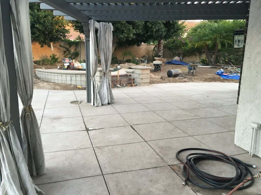 pergola with new concrete patio with lines to look like tiles every 4 feet, with raised patio with retaining wall and pillars in background