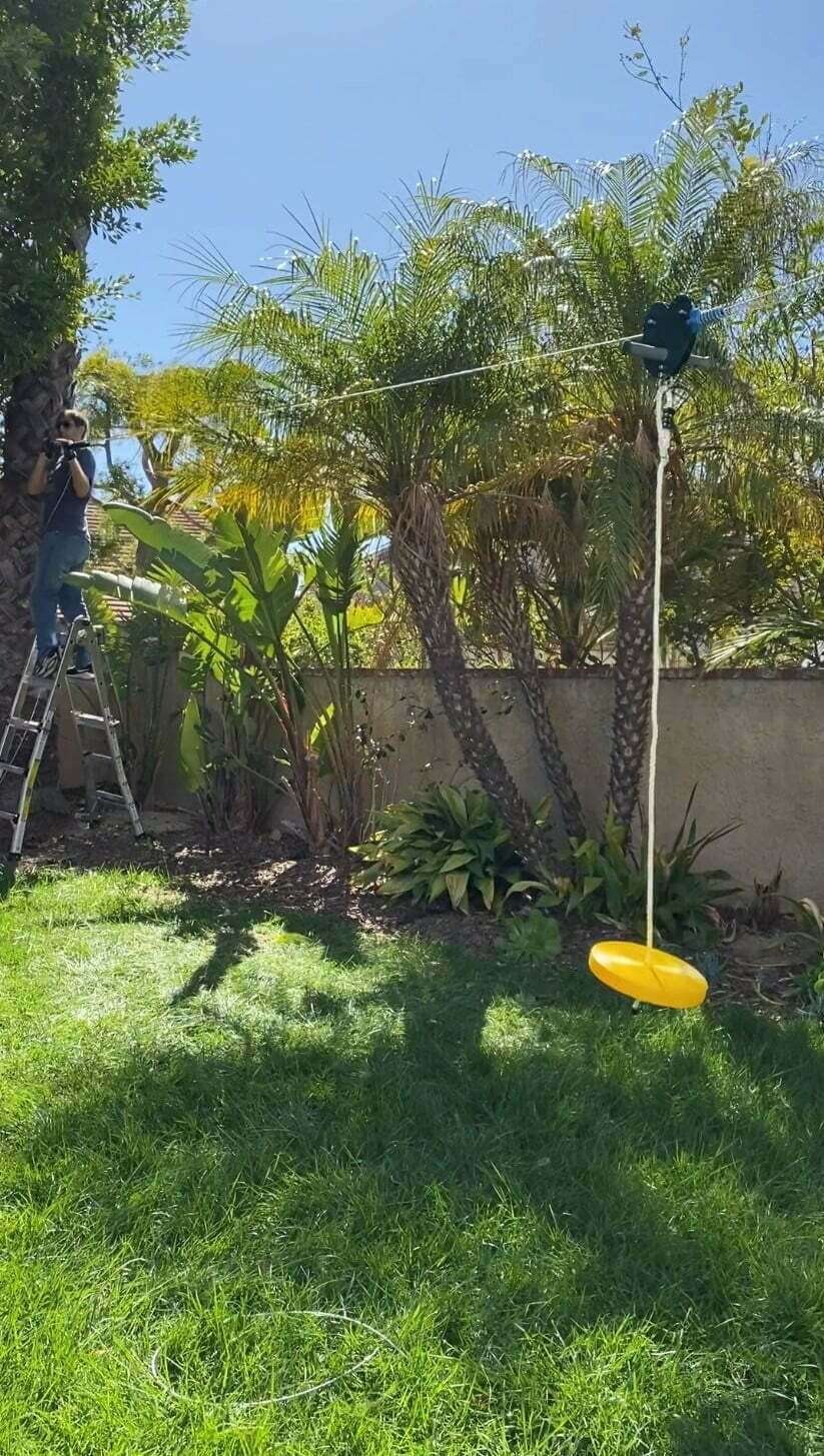 zipline lying on grass in backyard, with man installing zipline wire around tree in background