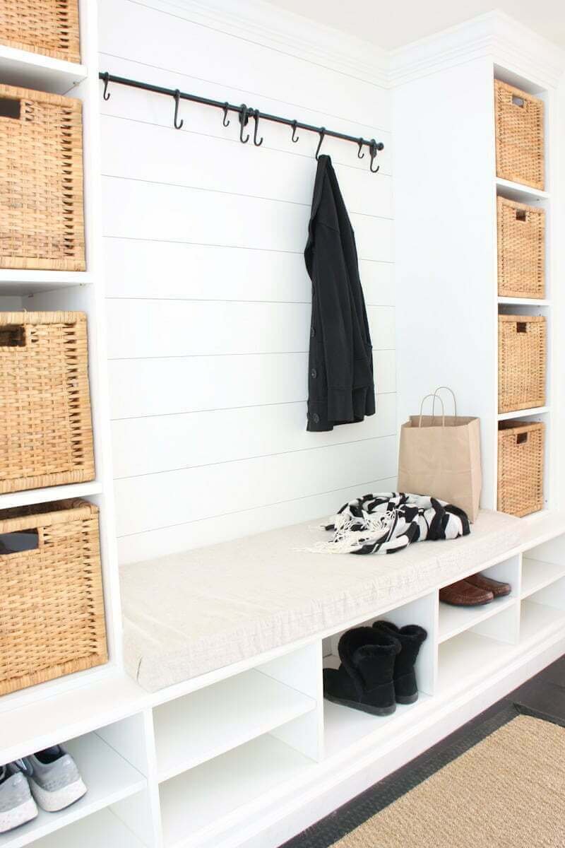 white mudroom with mudroom bench, shoe shelves, bench seat cushion, square baskets in cubbies, and black coat hooks on wall