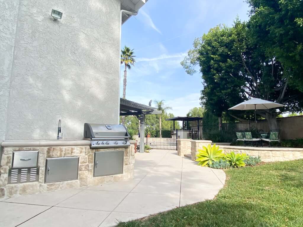 Backyard kitchen with outdoor kitchen grill, small outdoor kitchen sink, and stainless steel outdoor cabinets with view of patio dining area and pool in background