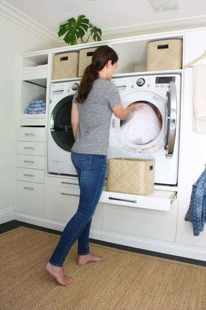 white laundry room with woman doing laundry pulling towels out of dryer with laundry baskets, shelves and drawers