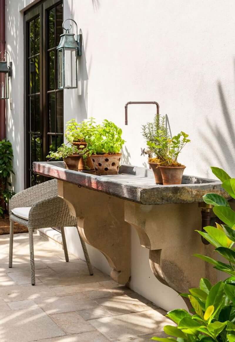 stone outdoor garden sink with brass faucet and corbels supporting, with pots of plants alongside house