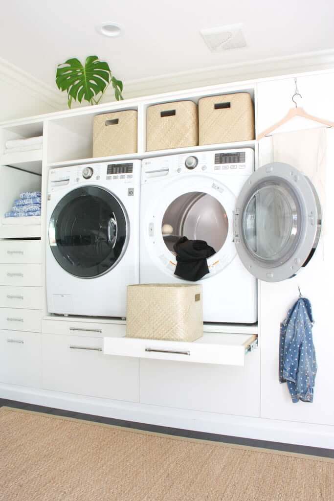 white laundry room decor with woven baskets, wooden hangers, open shelving, plants and drawers, with dryer open and basket on shelf for clean laundry