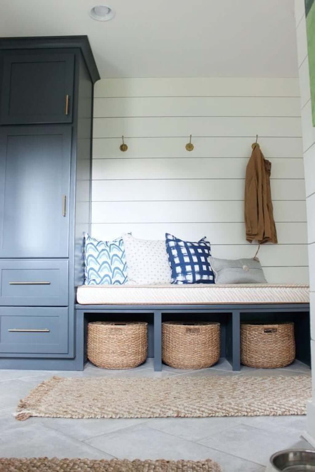 white and grey small mudroom bench with cushion, shiplap walls, round baskets for shoe storage, woven rug, and gold hooks with pillows