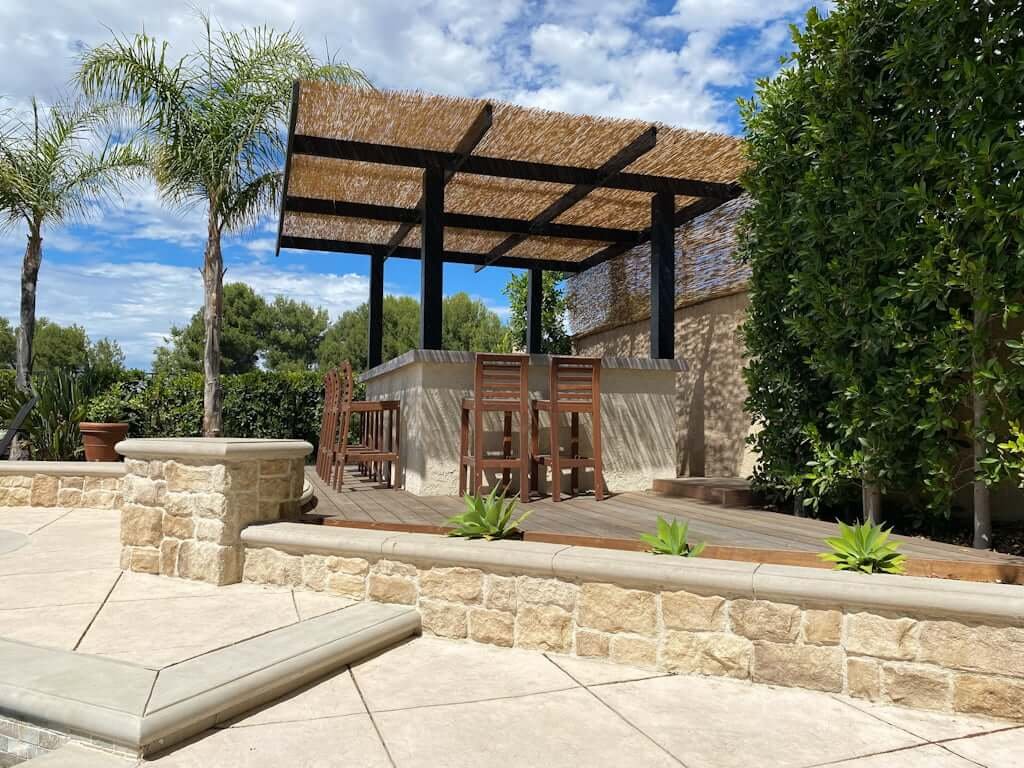 backyard bar with outdoor barstools, stucco wall, concrete counter top, and black pergola with thatch cover near pool with concrete stone walls and decking