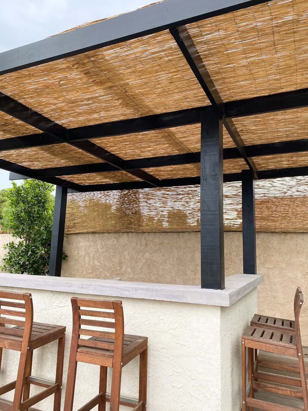 closet up outdoor bar with stucco wall, concrete counter top, wood patio bar stools, and black pergola with thatch cover near pool with concrete stone walls and decking