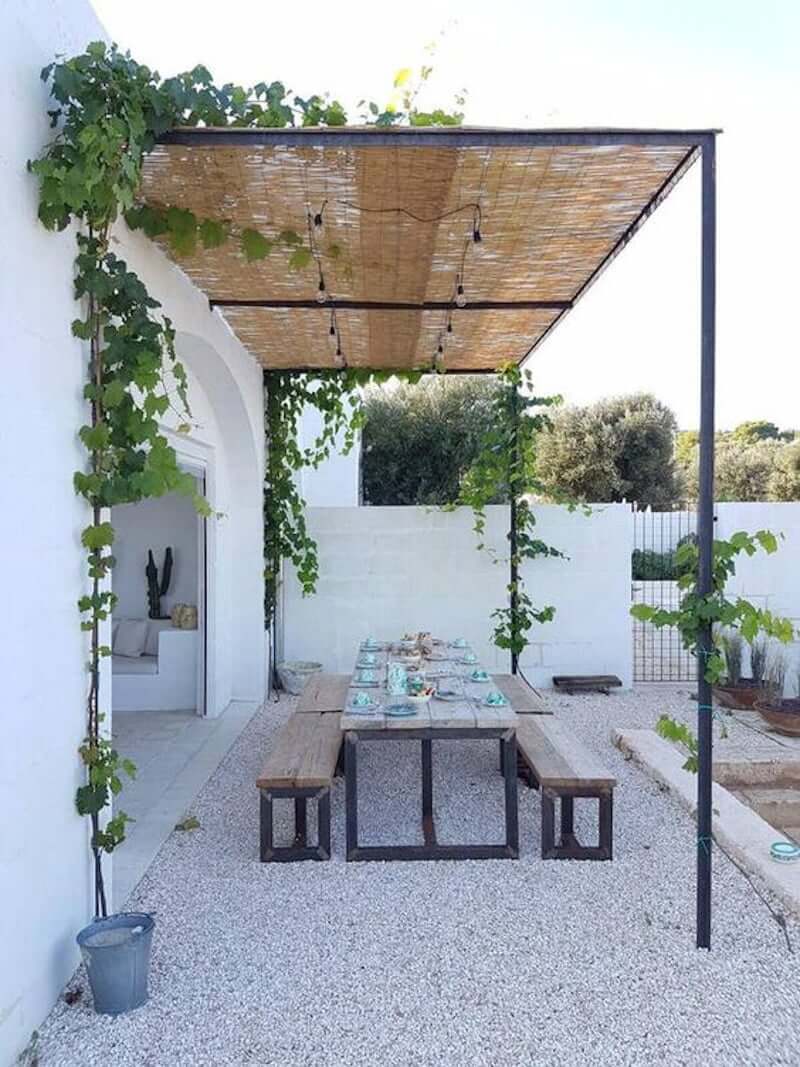 pea gravel dining area with table and benches, covered with thin thatch and black metal pergola cover extending over it