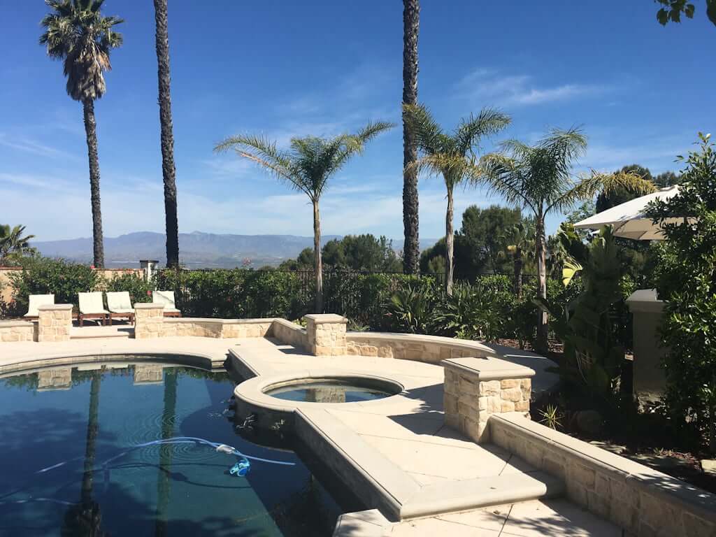 backyard pool and jacuzzi with concrete, stone veneer walls, patio bar to side, palm trees, and mountains in distance
