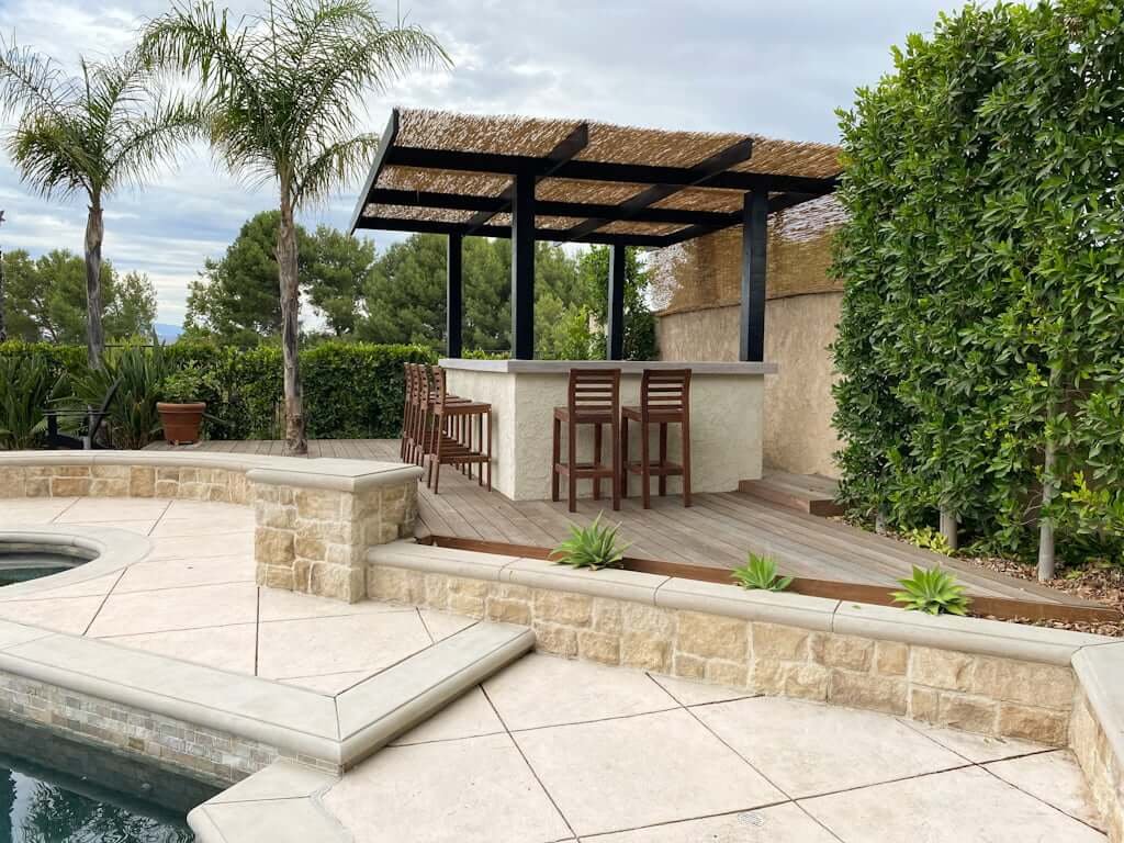 outdoor patio bar with concrete countertop, patio bar stools, and pergola with thatch cover near pool with concrete stone walls and decking and palm trees