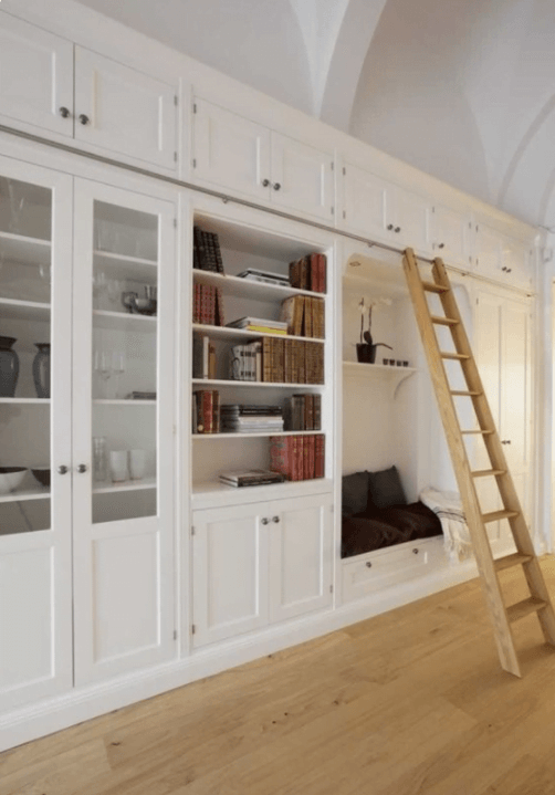 white bookshelves with glass doors, seat with cushions and pillows, and wood library ladder slanted with rail