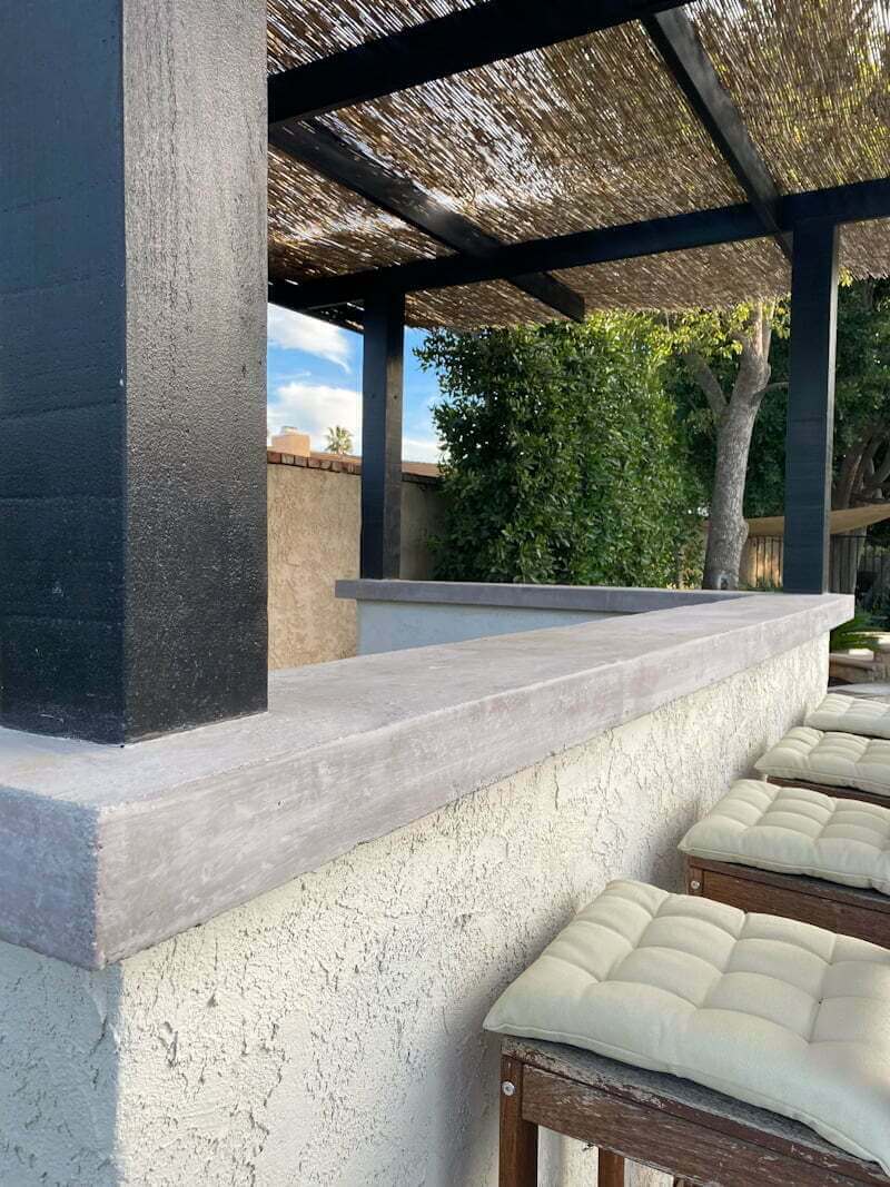 DIY concrete countertops in outdoor bar with stucco wall and bar stools and pergola with thatch roof
