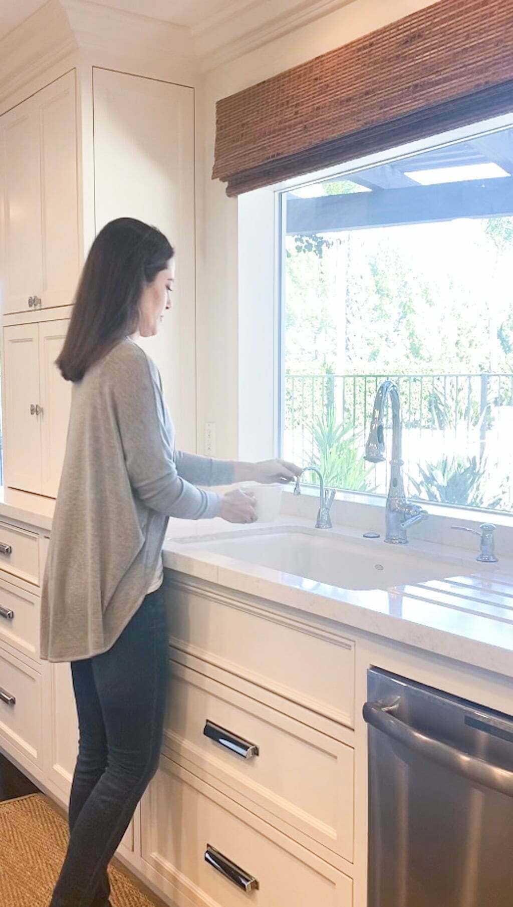 woman using instant hot water dispenser at sink in white kitchen with large picture window looking out to backyard