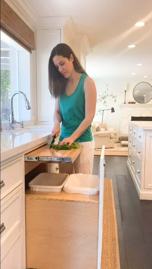 woman in white kitchen chopping food into pull out trash can