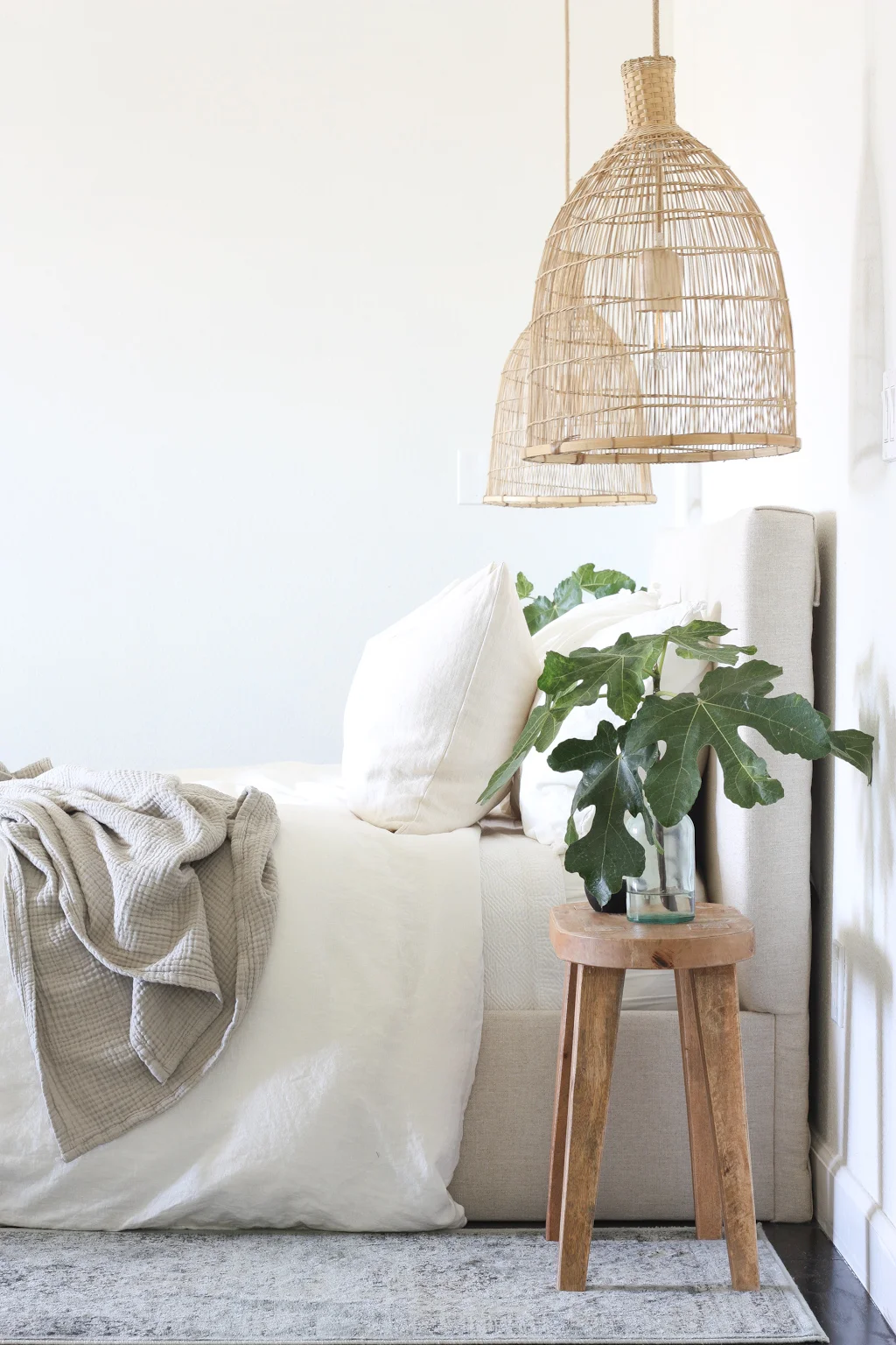 Guest bedroom decor cozy. Woven lights hang down on each side of the upholstered bed. Wooden nightstand with a vase full of greenery sit on top of the nightstand.