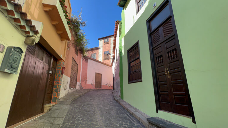 Small street in Canary Islands with green house then pink house on your right and green house, orange house and then pink house on your left