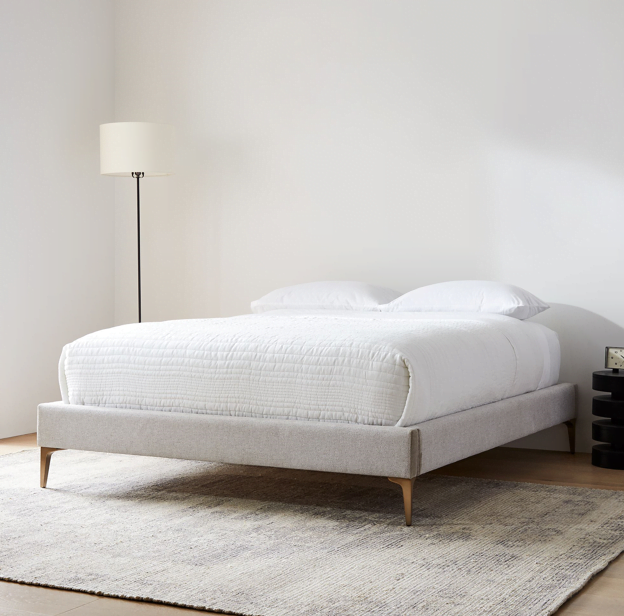 Upholstered bed frame design with no headboard sits on a rug in a room with white walls and one black floor lamp.