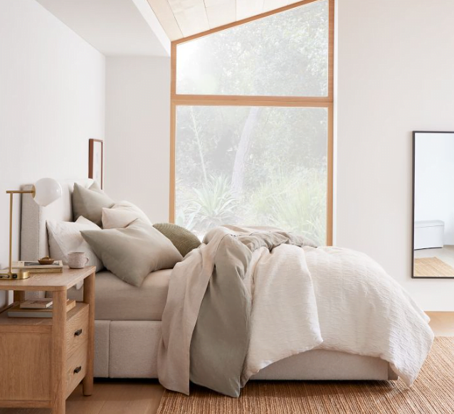 Side view of an upholstered bedframe with storage and neutral bedding. Wooden nightstand and jute rug below the bed. Large wooden window on one wall.