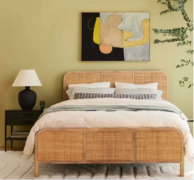 Woven bed frame with headboard and neutral bedding. One black nightstand sits to the side of the bed and one large piece of art hangs above the bed. 
