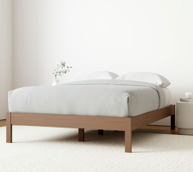 Minimalist wooden bed frame design in a room with white walls and a white rug below the bed. 