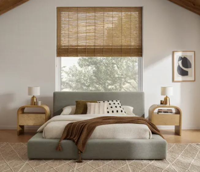Upholstered bed frame with storage that sits low to the ground with one large window behind the bed and a woven roman shade on the window. Two wooden nightstands on each side of the bed. 
