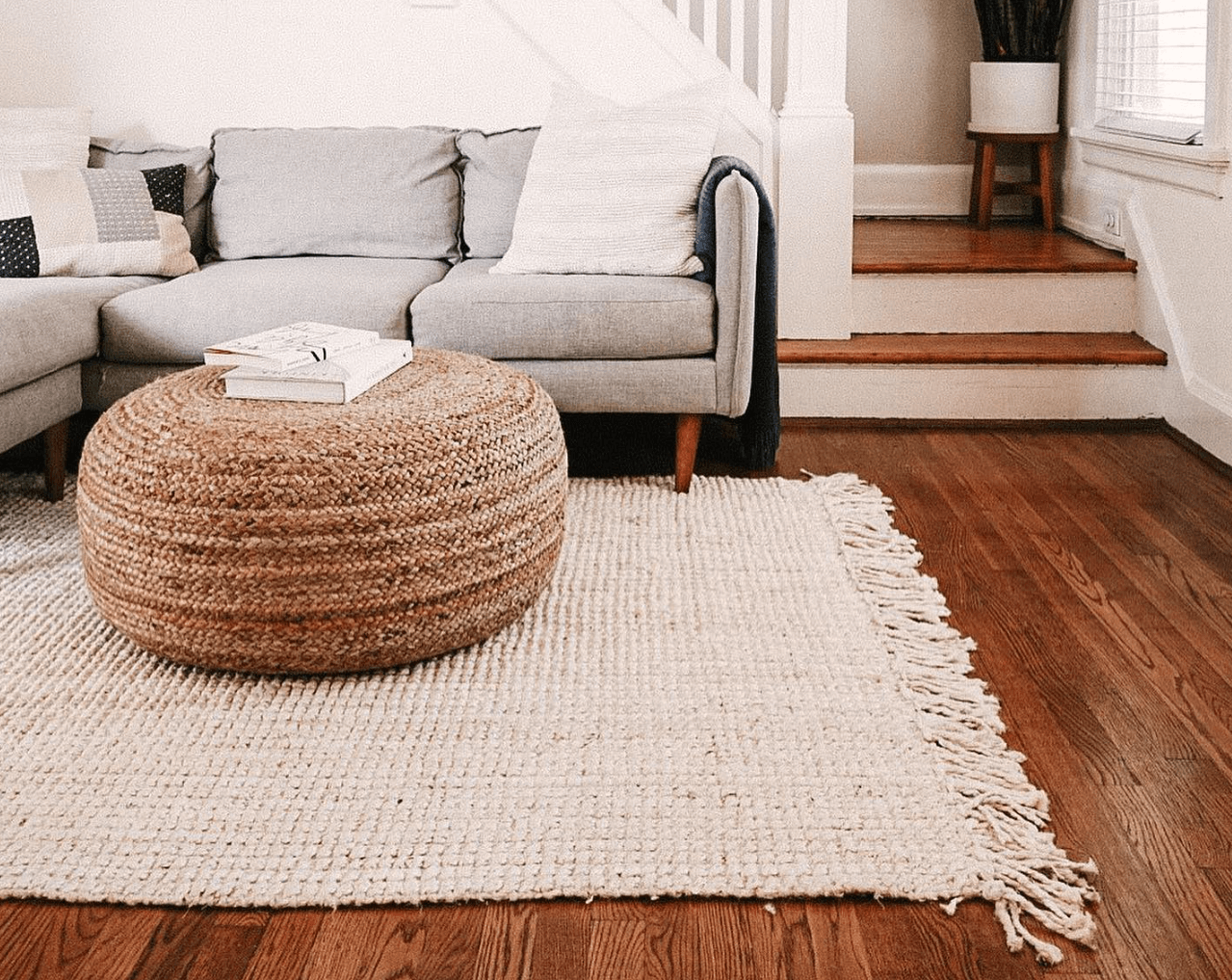 Living room with a light gray sofra and a woven coffee table with a white area rug with tassels on it. 