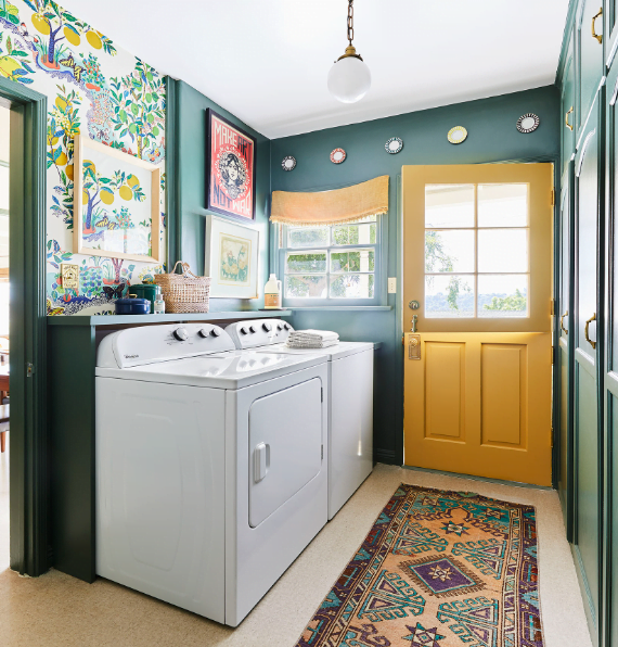 A dark green laundry room color with a yellow door and floral wallpaper on the walls. Plates and wall art hang on the walls. A shallow shelf is behind the top loading washer and dryer. 