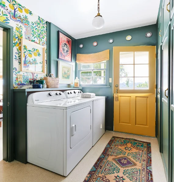 A dark green laundry room color with a yellow door and floral wallpaper on the walls. Plates and wall art hang on the walls. A shallow shelf is behind the top loading washer and dryer. 