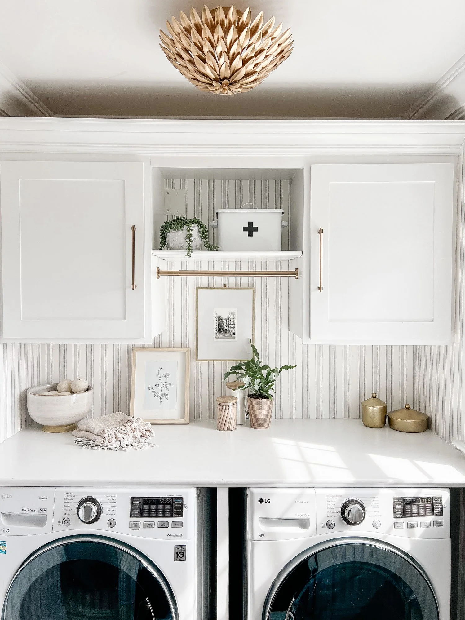 Striped laundry room wallpaper with white cabinetry and a statement gold light fixture. Laundry room art hangs on the wall. 