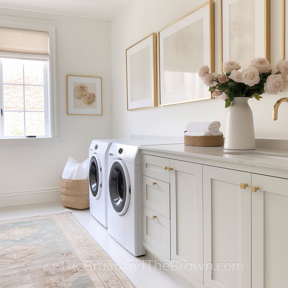 A light and airy laundry room decor ideas with white walls and laundry room art hanging on the walls. A woven basket sits on the floor. A vase full of flowers sits on the countertop. 