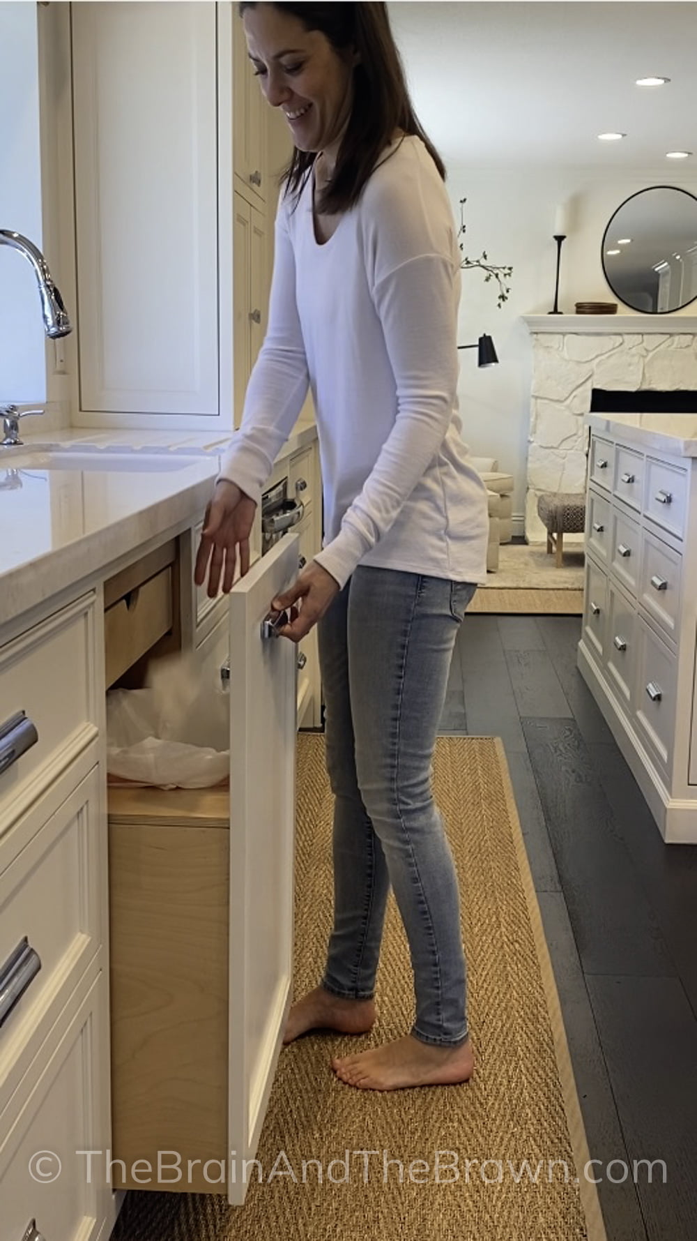 A women demonstrates how a pull-out trash drawer works in a spacious kitchen with white kitchen cabinets and white quartz countertops