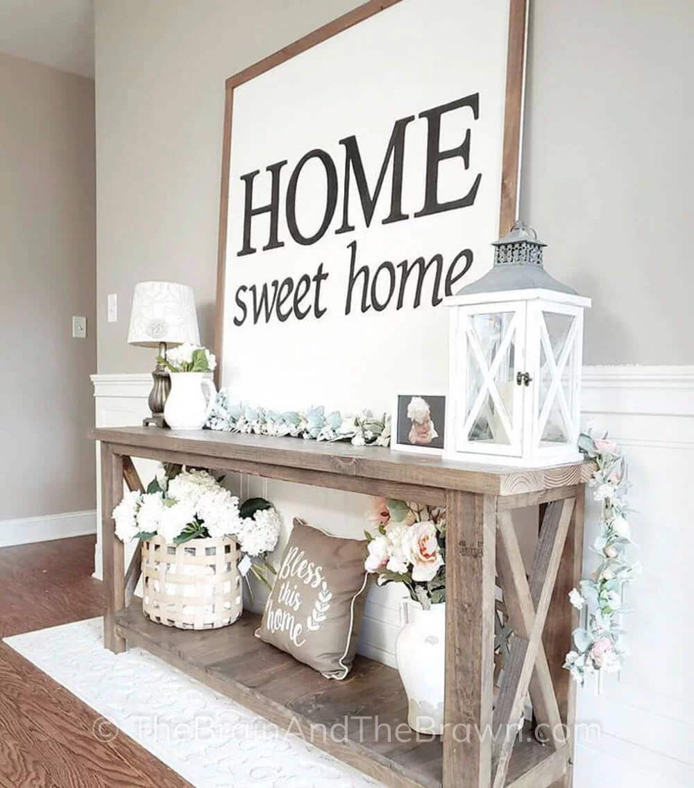 An entryway showcasing modern farmhouse ideas with a wooden console table and large sign leans on top of the console table. The console table is decorated with a lantern, lamp, flowers, baskets, picture frames and a pillow. 