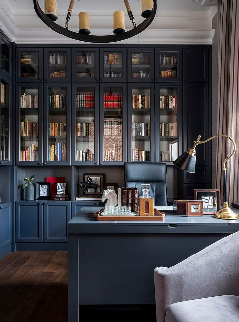 An office with dark painted floor to ceiling built in bookshelves with glass upper cabinets