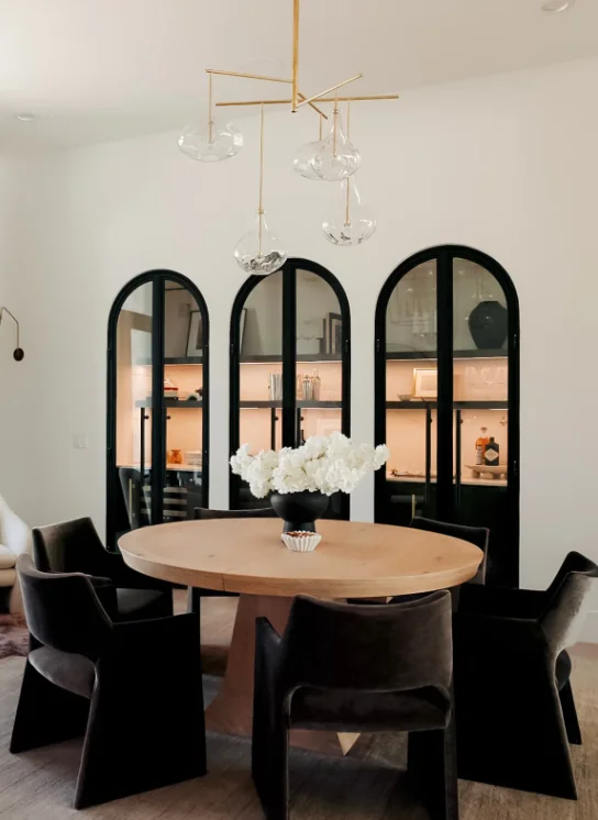 A dining room with a round wooden table and three arched built in cabinets with glass doors