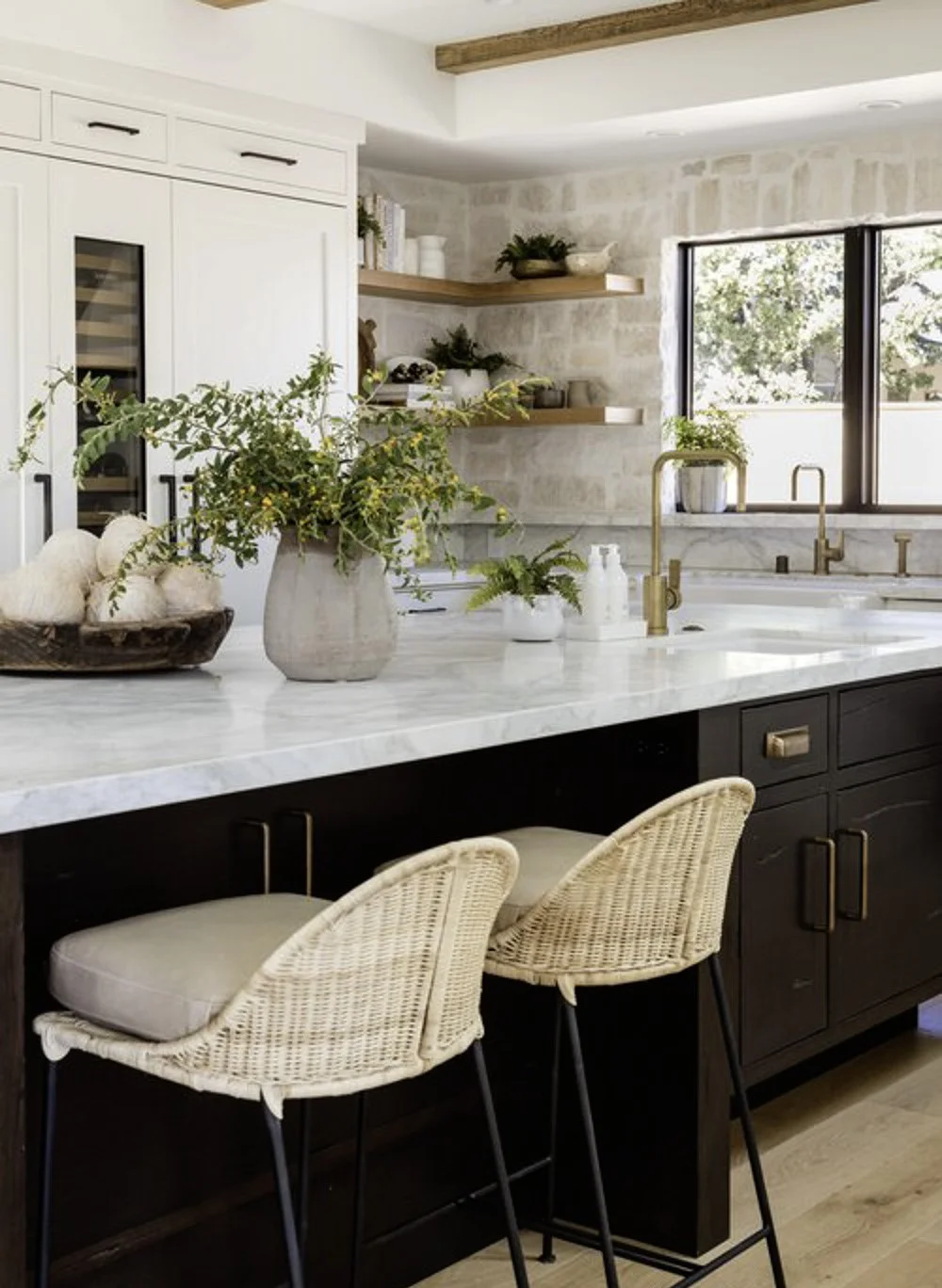 Over grouted stone backsplash, white perimeter kitchen cabinets, black island, marble countertops and brass hardware