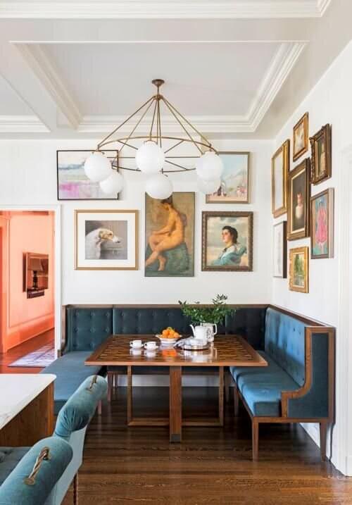 An eclectic dining space with a large gallery wall, blue velvet banquette seat and square wooden dining table 