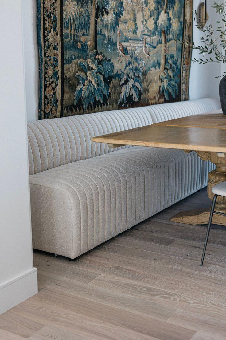 A cream upholstered bench with a large tapestry hanging above it and a wooden rectangular table in front of the bench