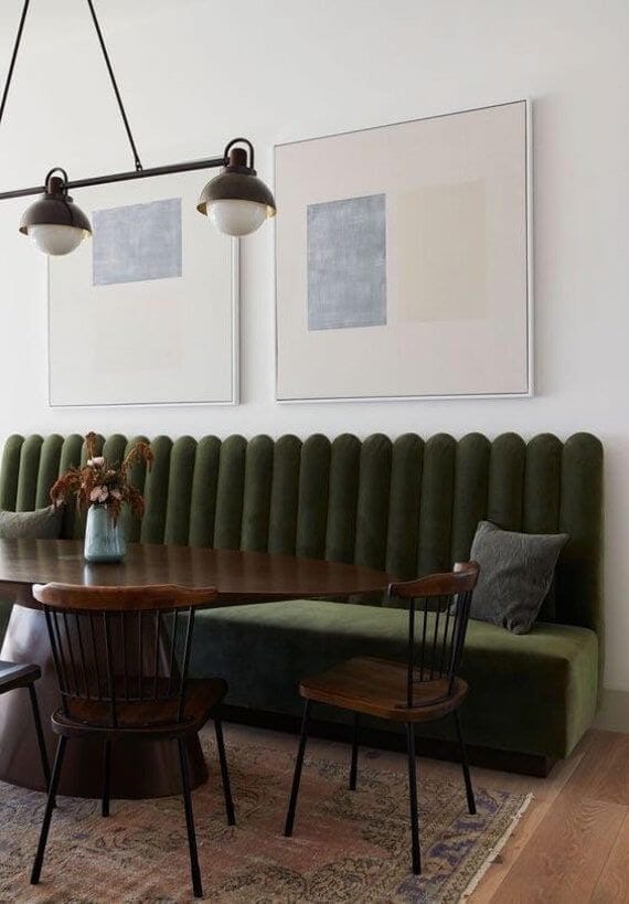 Green velvet channeled banquette bench against a wall with large artwork above it and an oval wooden table in front of it