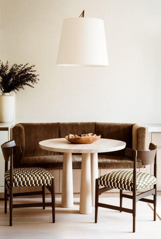 A small dining space with a brown velvet banquette seat, a white round stone table and two wooden chairs