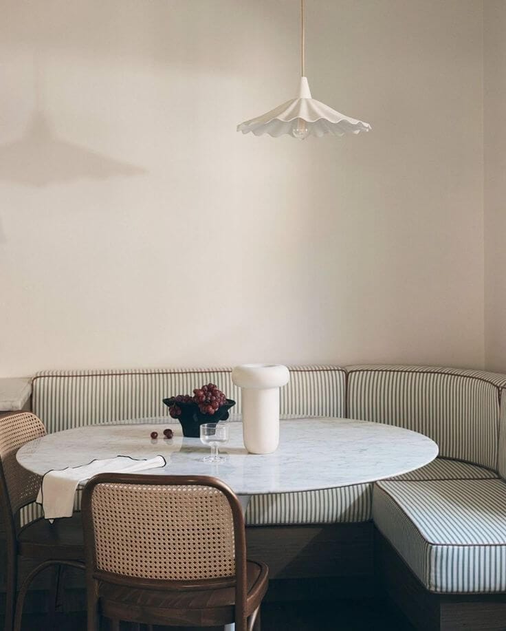 A dining room with a green striped curved banquette bench, a white tulip table and two wooden and cane chairs opposite the bench