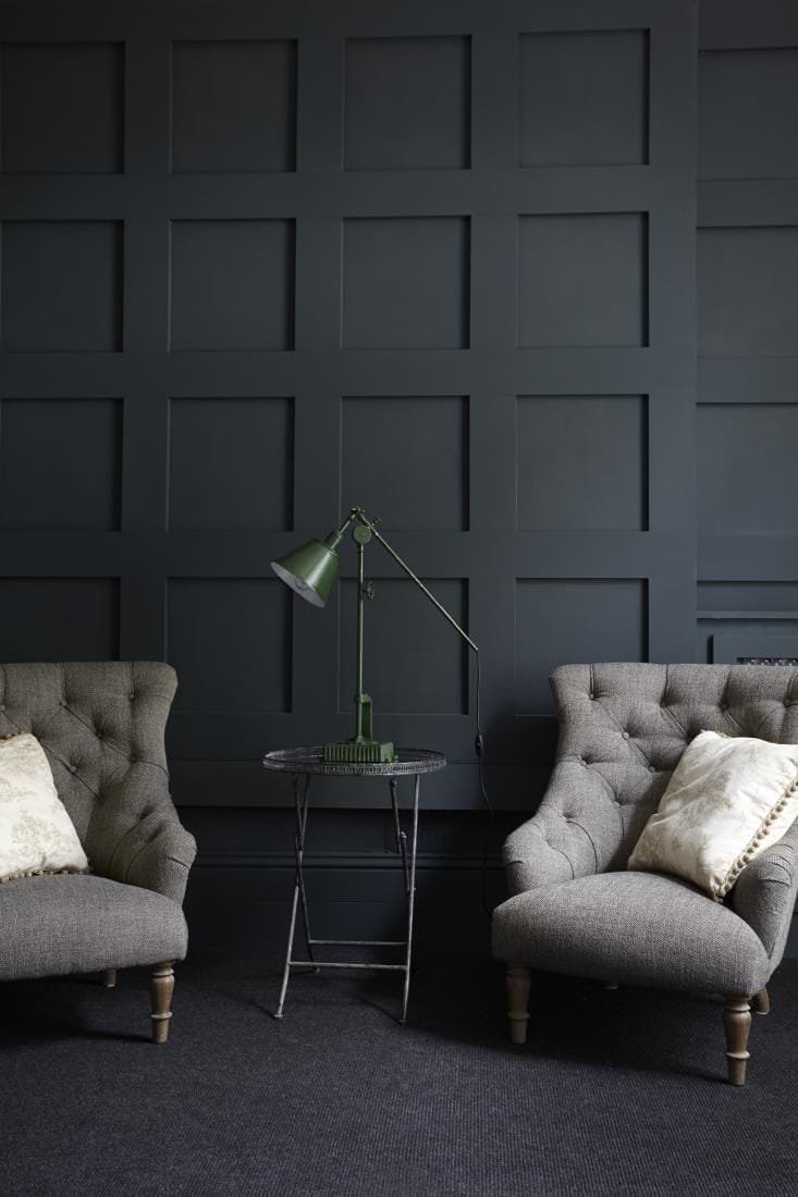 A dark and moody room with a square grid accent wall painted a dark charcoal color with two grey chairs 