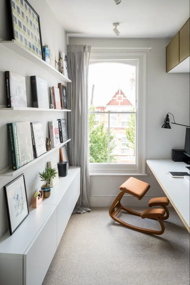 a narrow white home office with window and ergonomic chair, with open shelving and books on one side opposite a floating white desk
