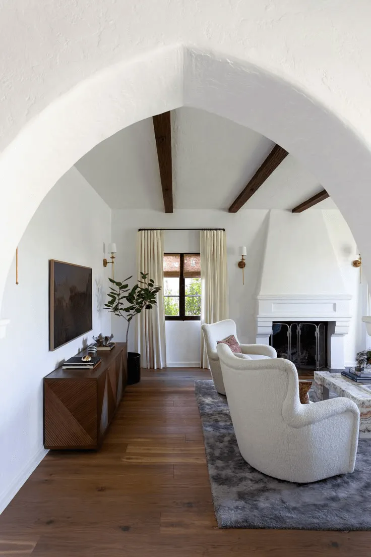 a Spanish-style house interior showing a living room with fireplace and TV, arched doorway and wood ceiling beams