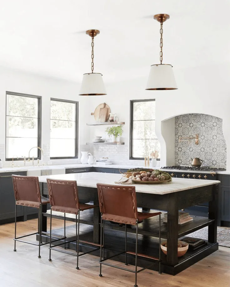 Spanish style kitchen interior with tile backsplash behind stove and a large island, leather chairs and ceiling pendants