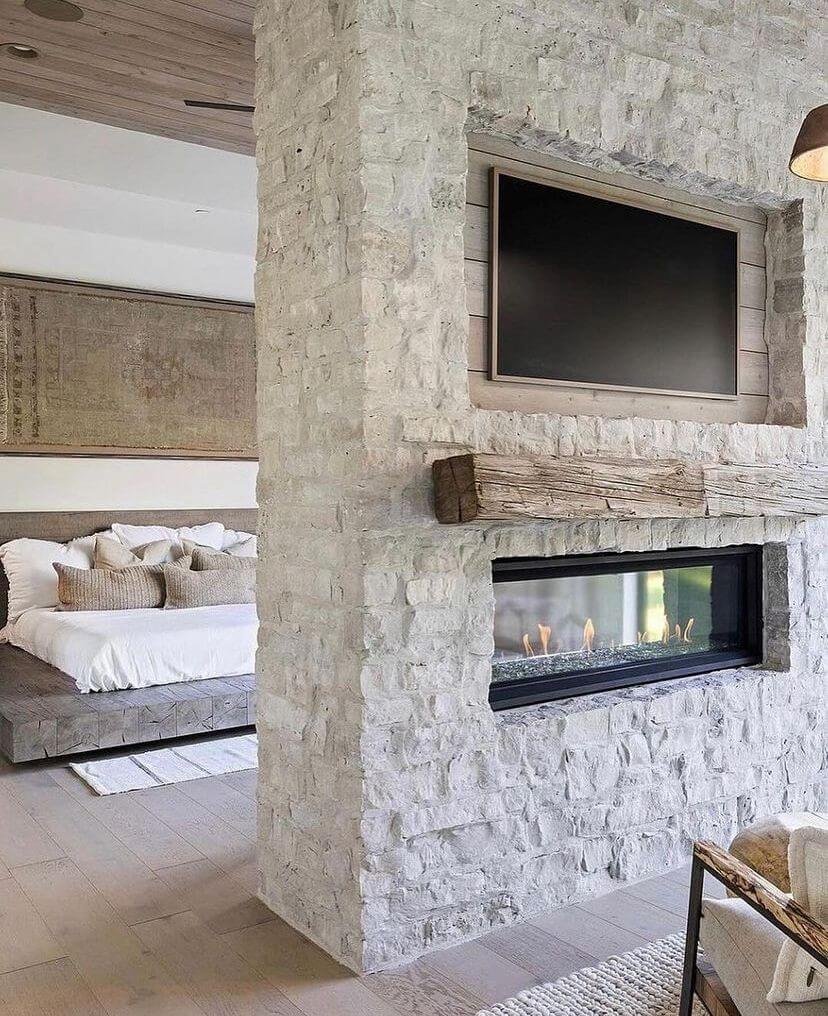 a double-sided stone fireplace in a bedroom with TV, natural wood mantel from reclaimed wood, and a gas fireplace